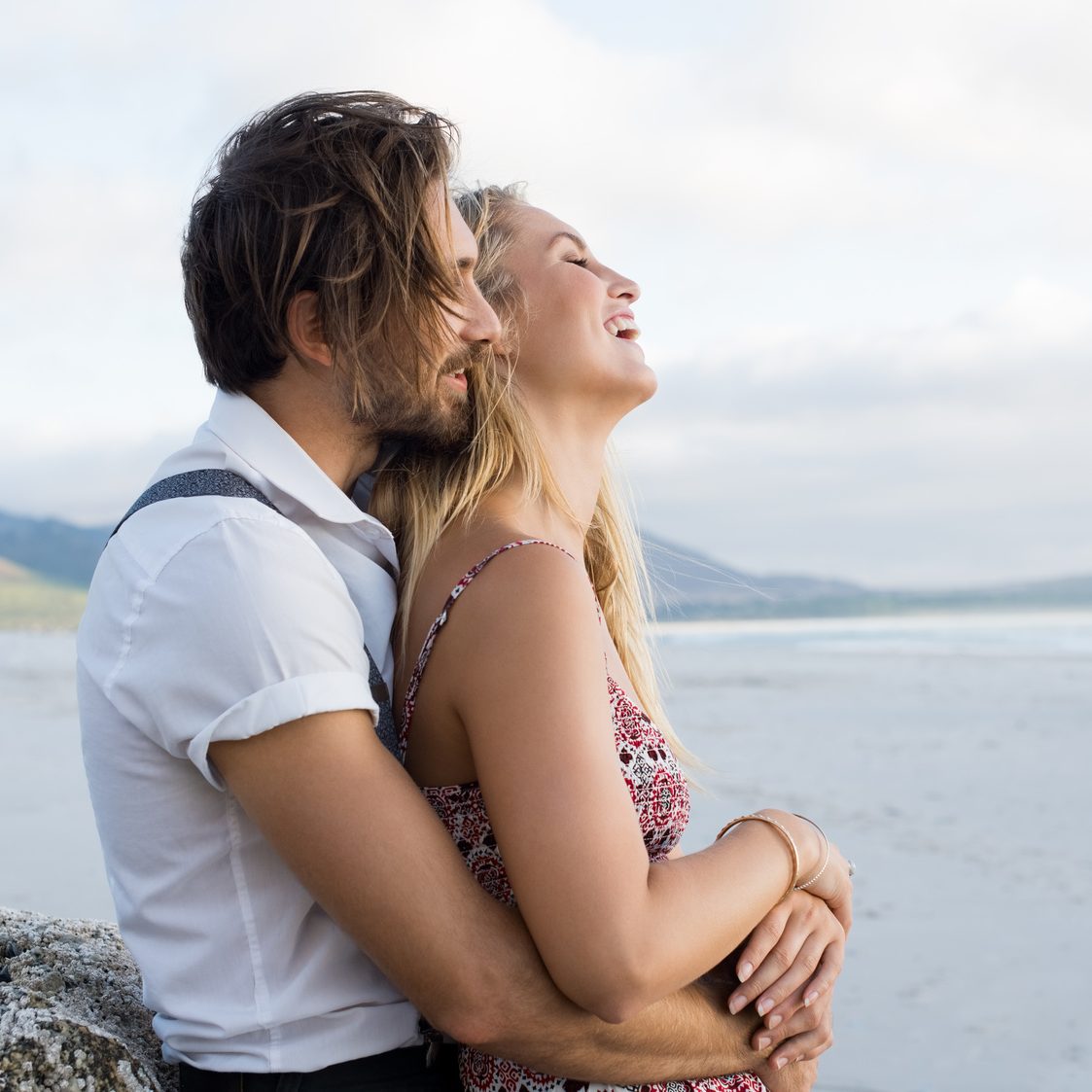 Couple hugging by the sea. Young couple at the beach holding each other. Boyfriend embracing girl from behind at beach during sunset at seashore.